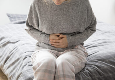 Sick woman in grey homewear sitting on bed, keeping hands on stomach, suffering from intense pain. Illness, stomach ache concept